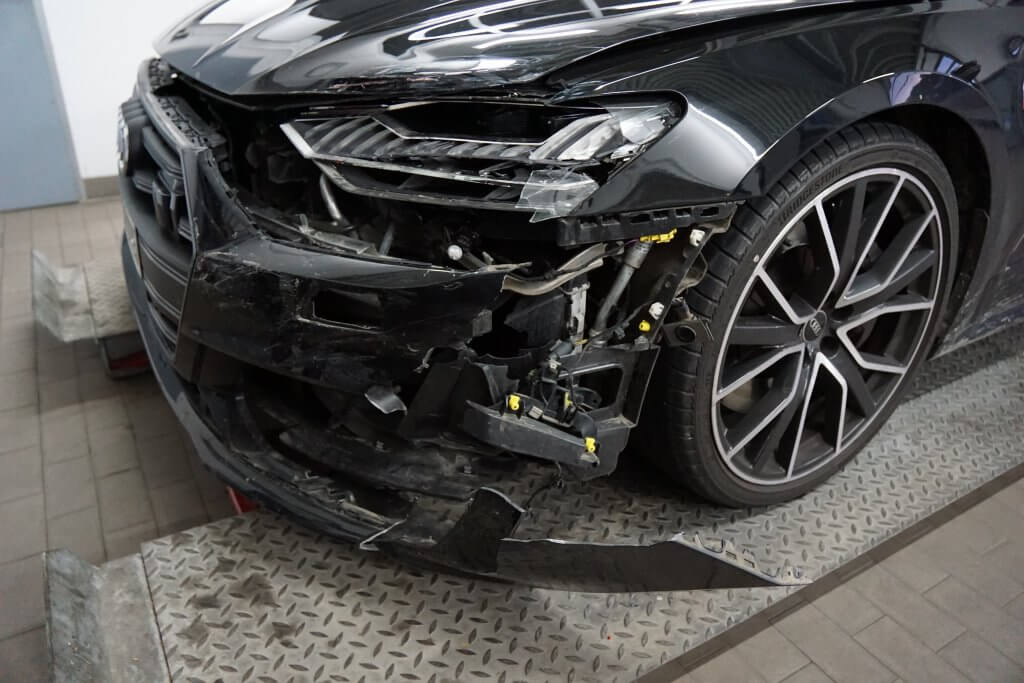 Ein schwarzes Auto mit stark beschädigter Front und kaputter Stoßstange steht auf einer Metallplattform in einer Werkstatt