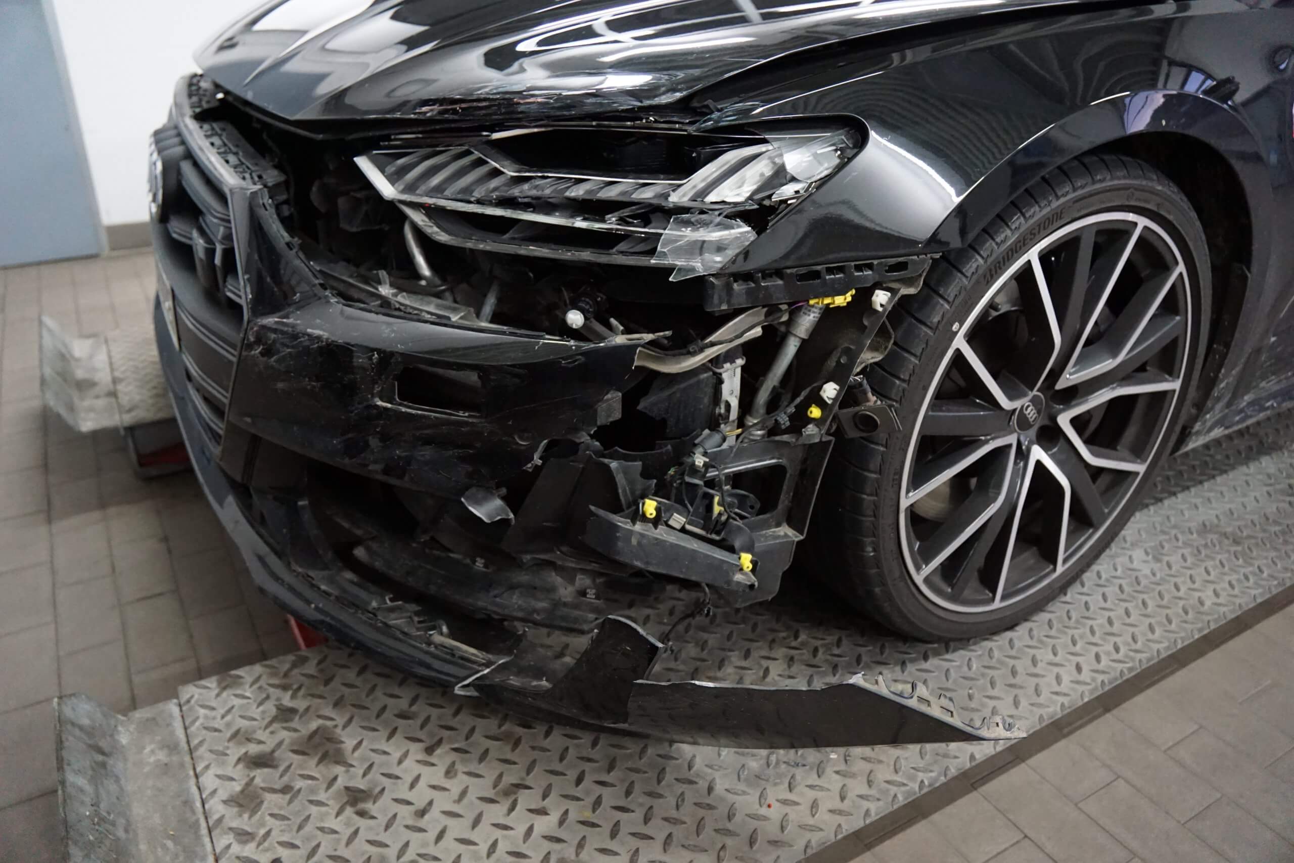 Ein schwarzes Auto mit stark beschädigter Front und kaputter Stoßstange steht auf einer Metallplattform in einer Werkstatt