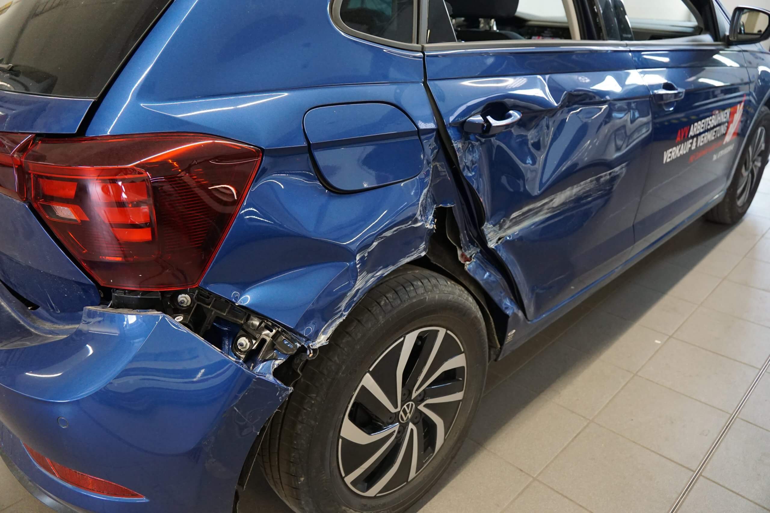 Ein blauer Wagen mit starken Schäden an der linken hinteren Seite, Dellen und verbogenem Radlauf, steht in einer Halle