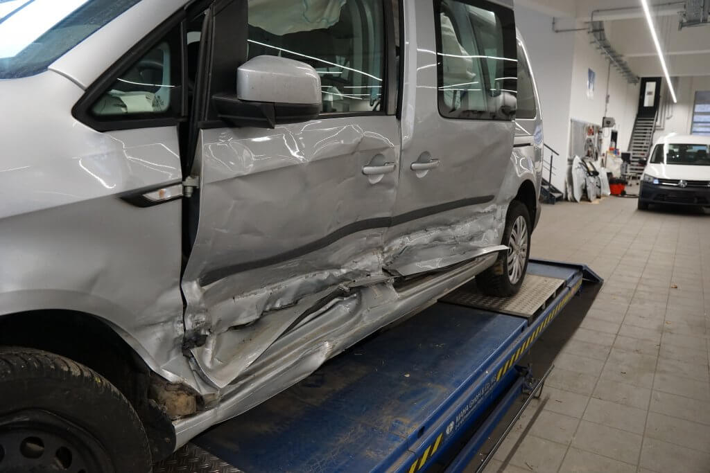 Ein silberner Van mit schweren Schäden an den Seitentüren steht auf einer Hebebühne in einer Autowerkstatt