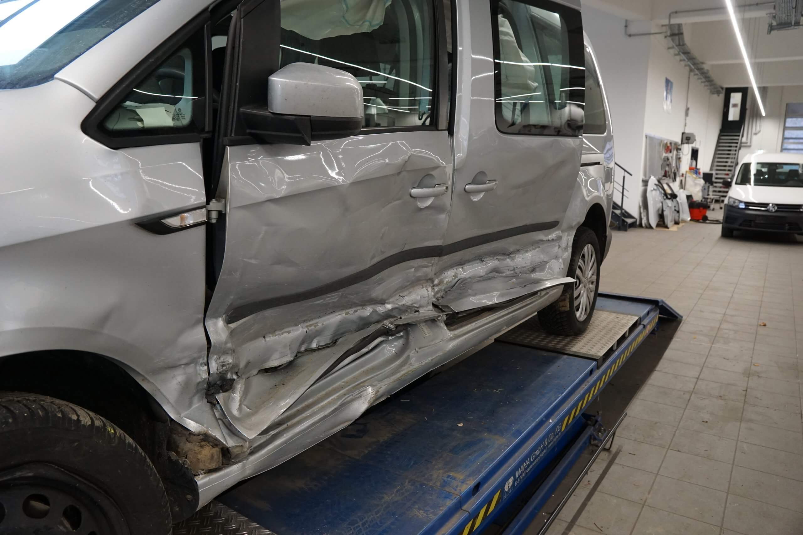 Ein silberner Van mit schweren Schäden an den Seitentüren steht auf einer Hebebühne in einer Autowerkstatt