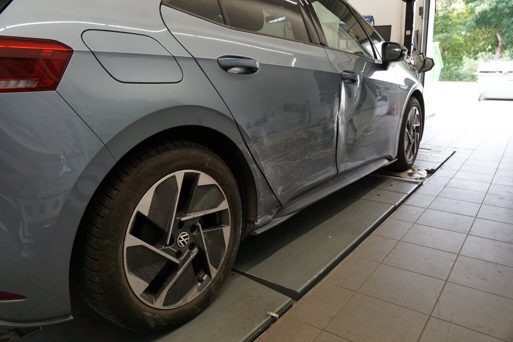 Ein graues Auto mit beschädigter Seitentür und Kratzern steht in einer Werkstatt oder Garage