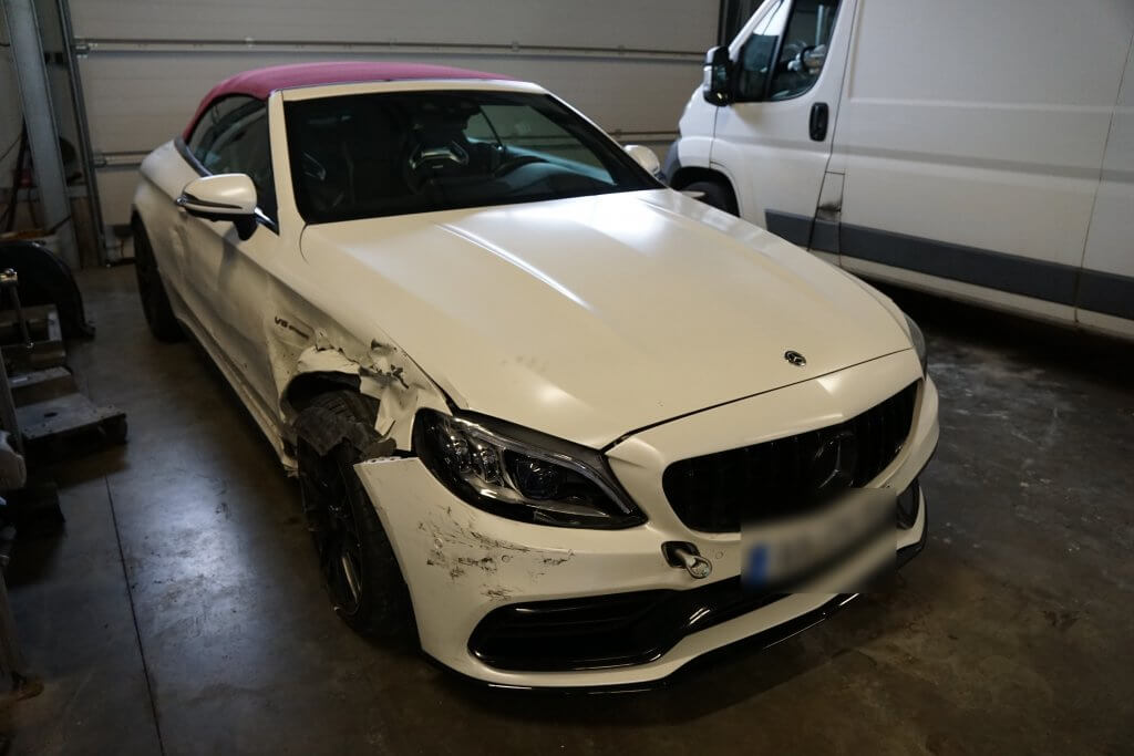 Ein weißes Mercedes Cabrio mit beschädigter Front links steht in einer Halle neben einem weißen Transporter