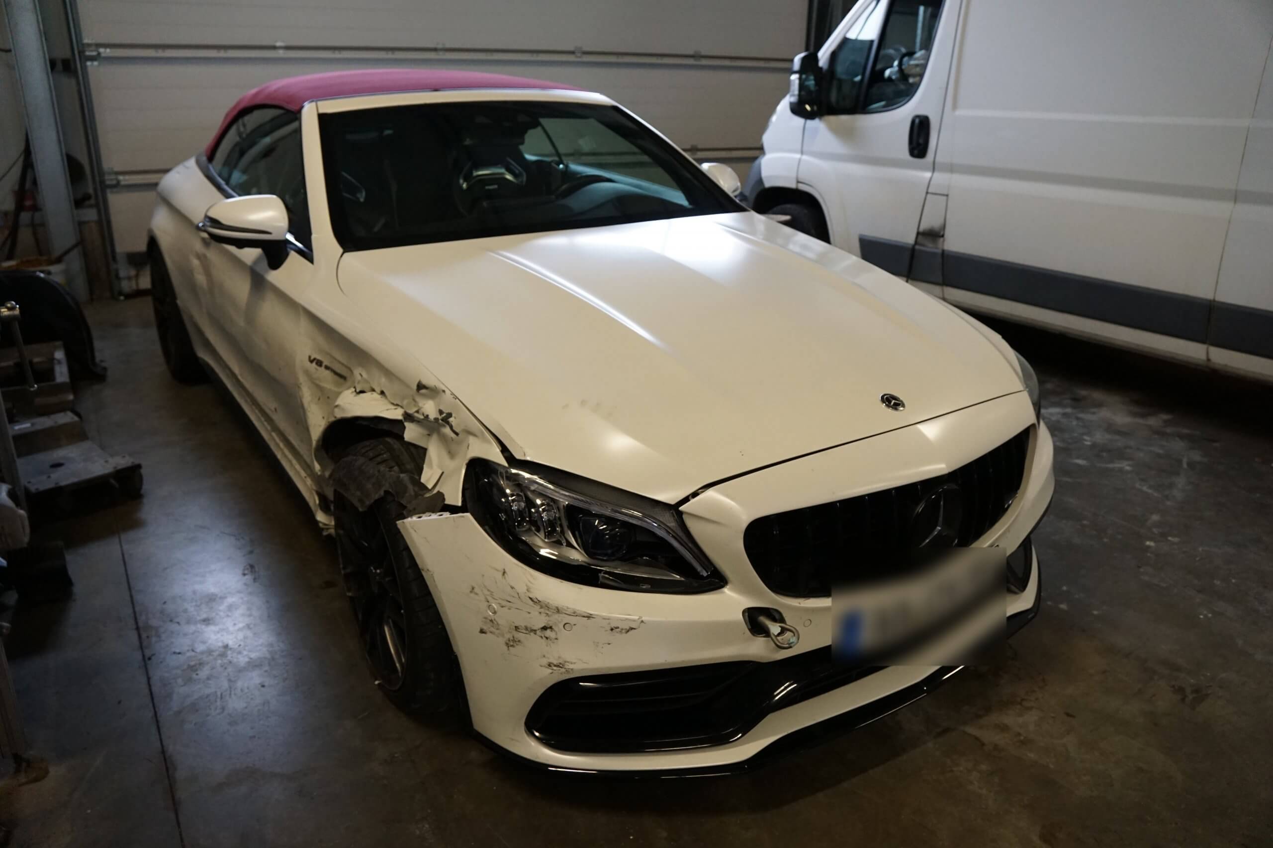 Ein weißes Mercedes Cabrio mit beschädigter Front links steht in einer Halle neben einem weißen Transporter