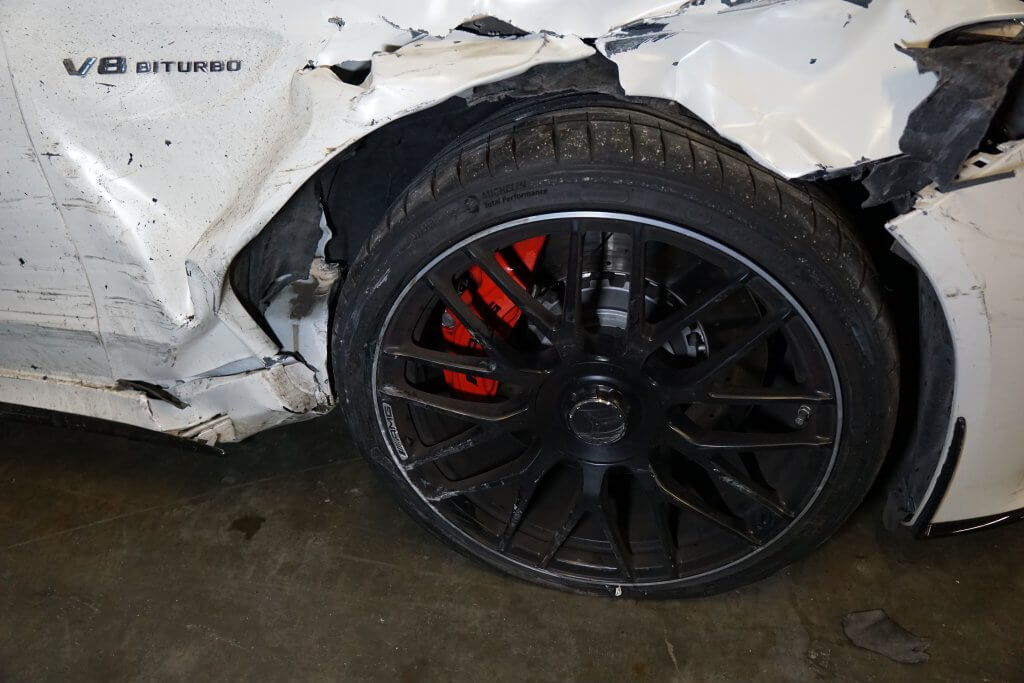 Nahaufnahme eines beschädigten weißen Autos mit eingedrücktem Kotflügel, verkratztem schwarzen Rad und V8 BITURBO-Emblem