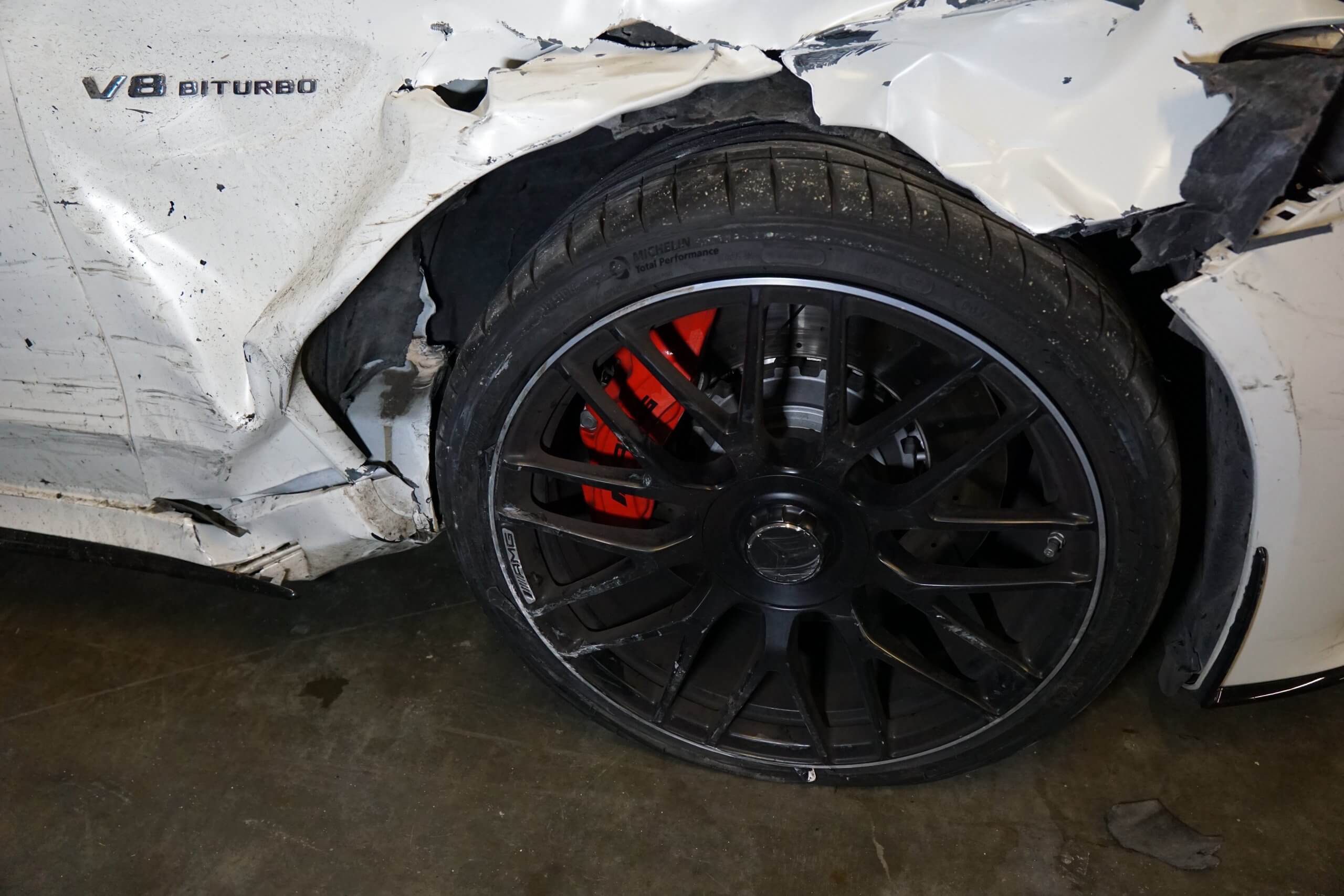 Nahaufnahme eines beschädigten weißen Autos mit eingedrücktem Kotflügel, verkratztem schwarzen Rad und V8 BITURBO-Emblem