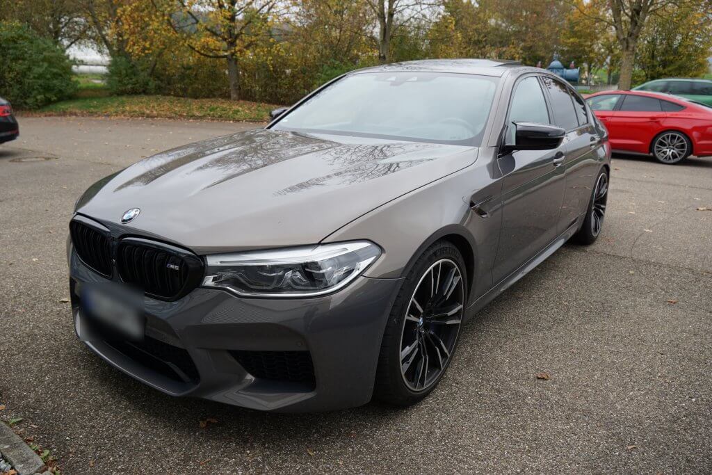 Ein grauer BMW steht an einem bewölkten Tag auf einem Parkplatz, umgeben von Bäumen und weiteren Autos