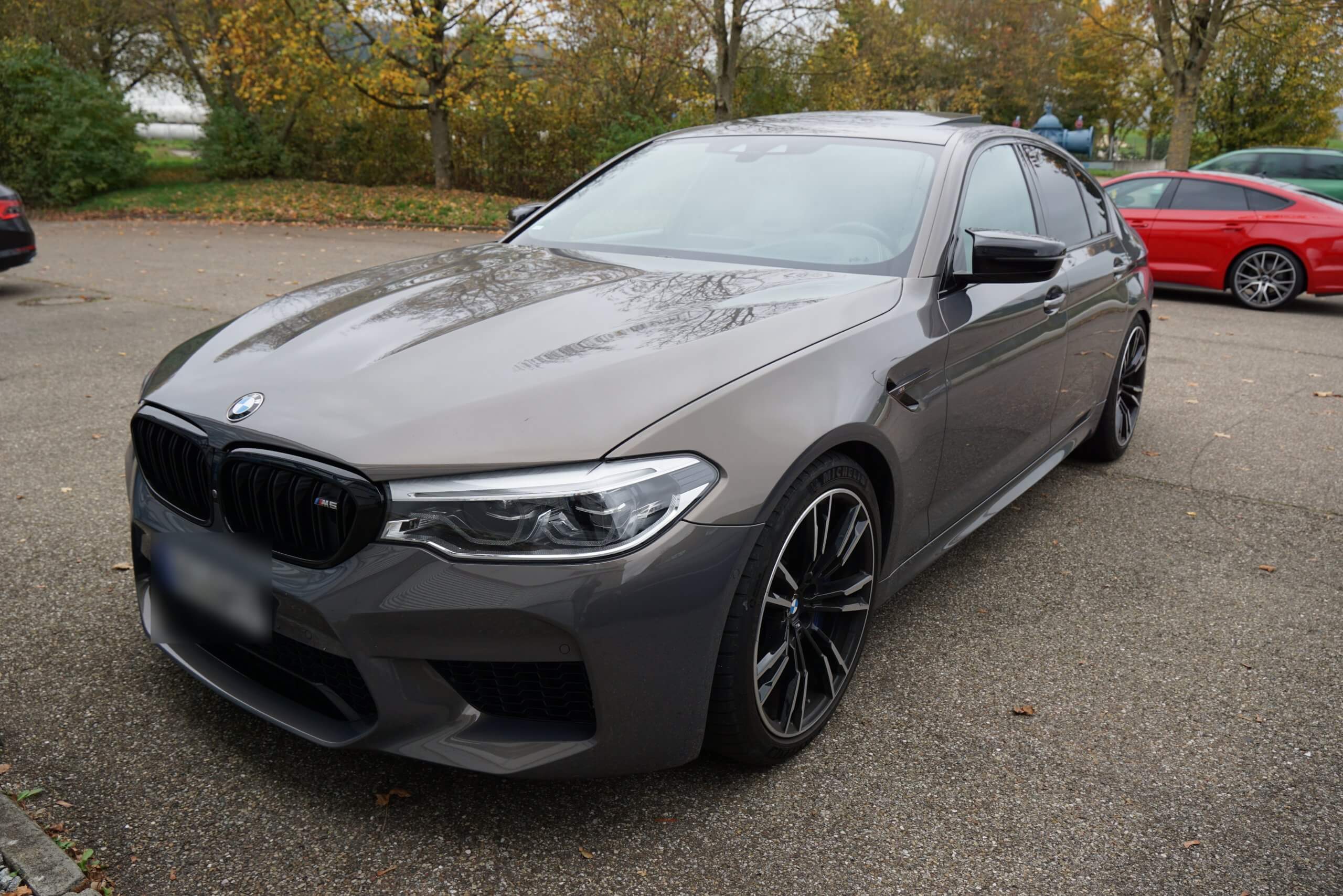 Ein grauer BMW steht an einem bewölkten Tag auf einem Parkplatz, umgeben von Bäumen und weiteren Autos