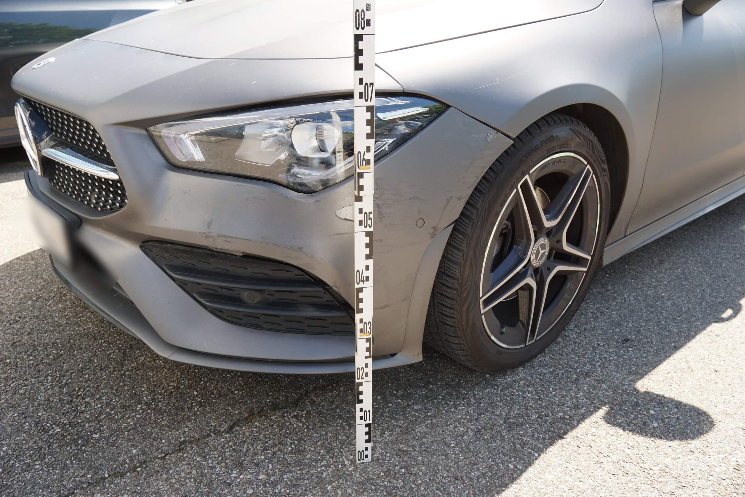 Ein Messstab steht vor Stoßstange und Rad eines grauen Autos auf Asphalt bereit für das Unfallgutachten