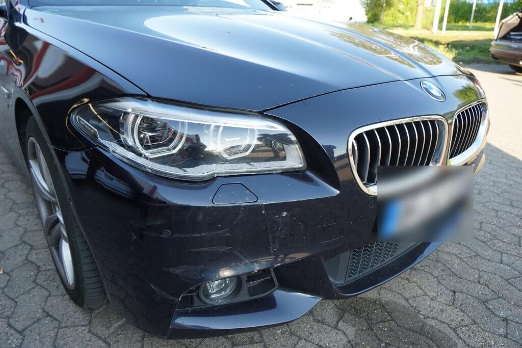 Nahaufnahme der dunkelblauen BMW-Frontstoßstange und Scheinwerfer auf sonnenbeschienener Straße, Nummernschild unscharf