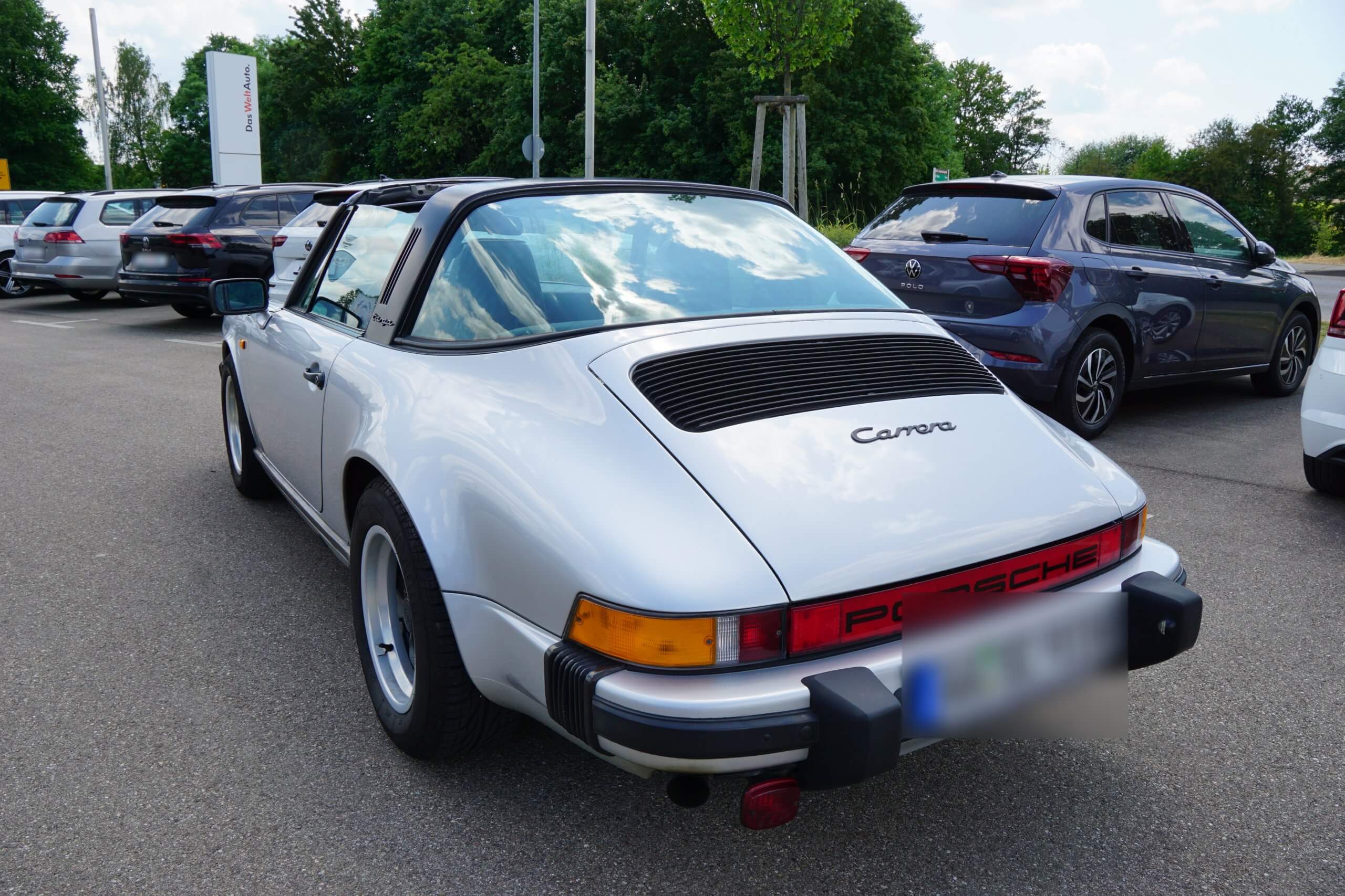 Ein silberner Porsche Carrera Targa steht an einem sonnigen Tag auf einem Parkplatz zwischen modernen Autos