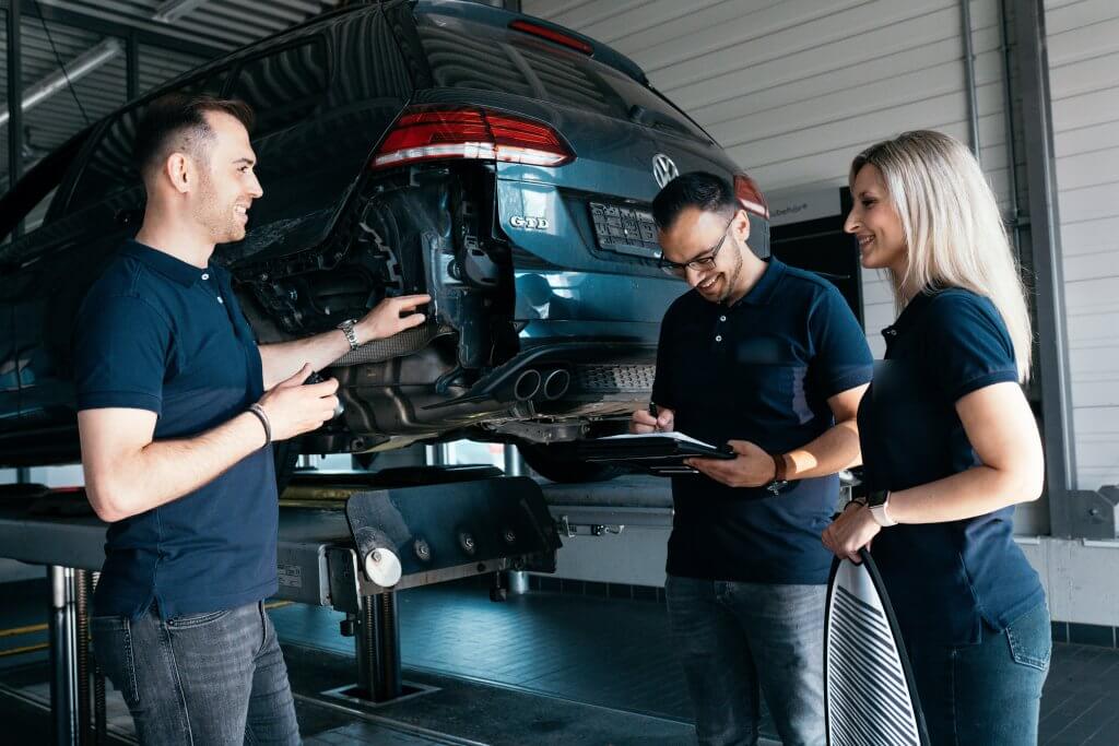 Drei Kfz-Mechaniker in dunkelblauen Hemden besprechen Reparaturen an einem Auto auf der Hebebühne, einer hält ein Klemmbrett