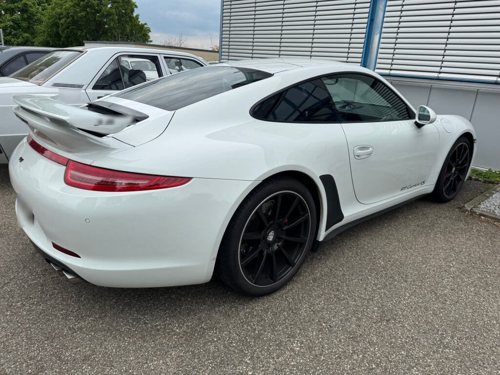 Ein weißer Porsche 911 mit schwarzen Felgen steht auf einem asphaltierten Platz neben einem weiteren Auto