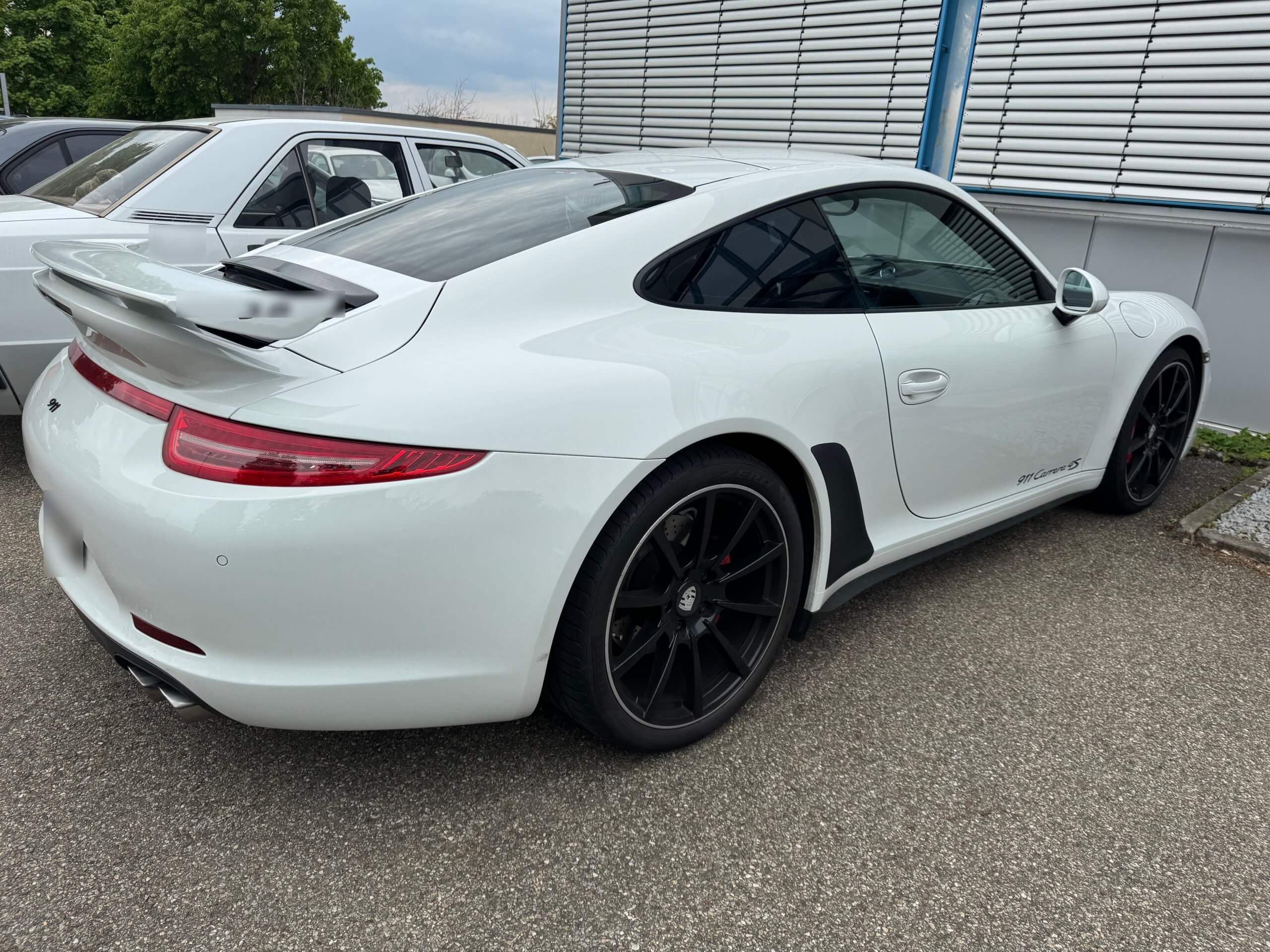 Ein weißer Porsche 911 mit schwarzen Felgen steht auf einem asphaltierten Platz neben einem weiteren Auto