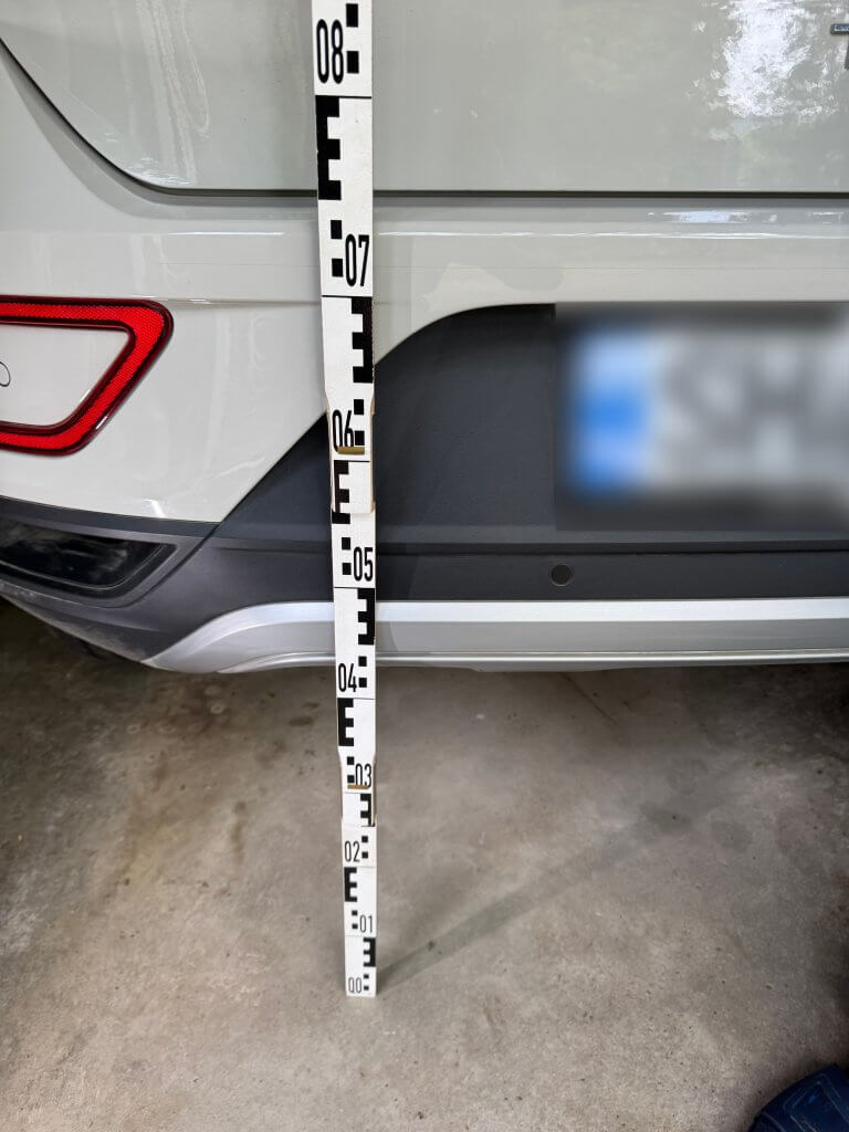 Ein Messstab wird senkrecht neben die hintere Stoßstange eines weißen Autos in einer Garage gehalten, das Nummernschild ist unscharf