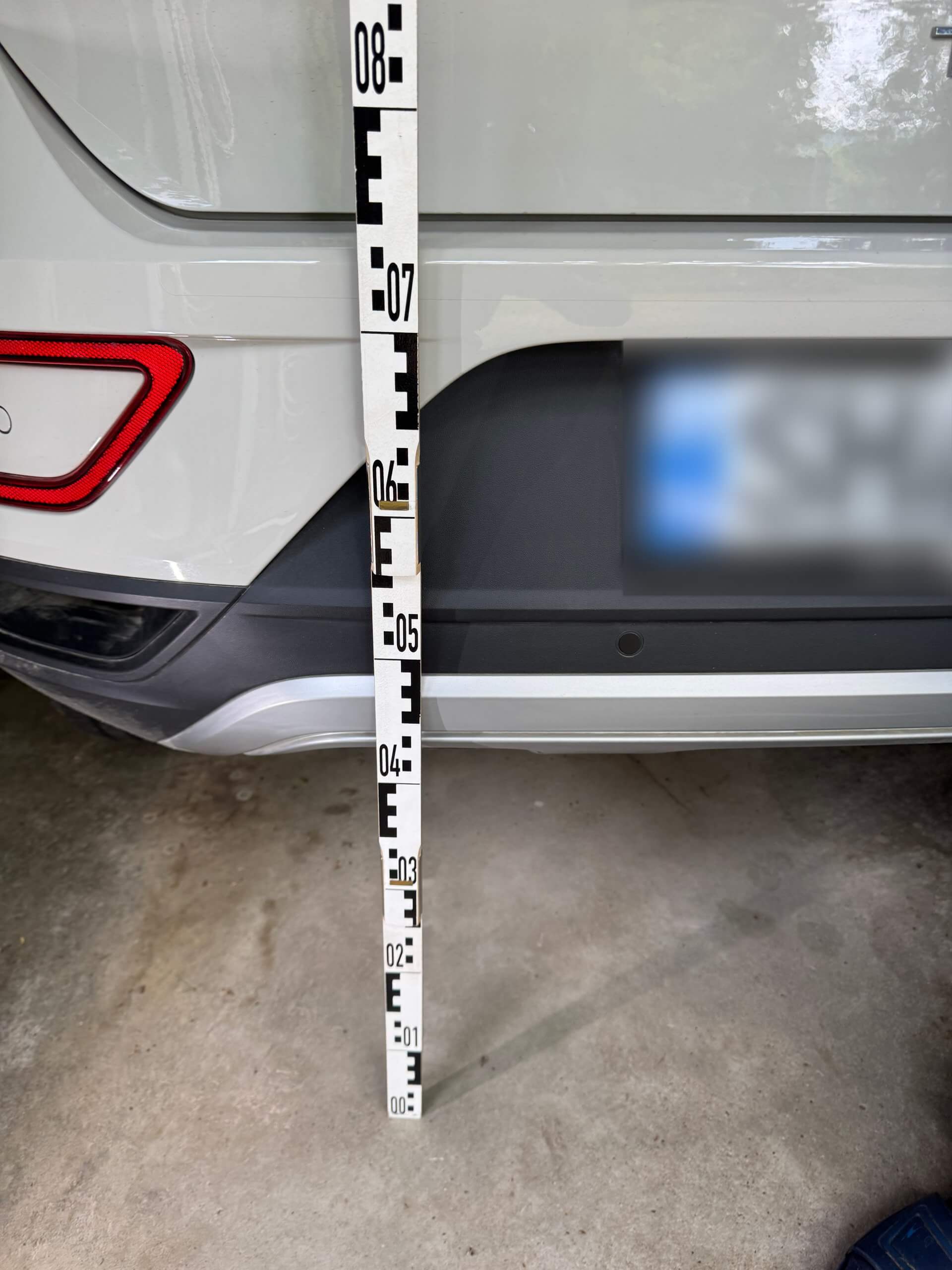 Ein Messstab wird senkrecht neben die hintere Stoßstange eines weißen Autos in einer Garage gehalten, das Nummernschild ist unscharf