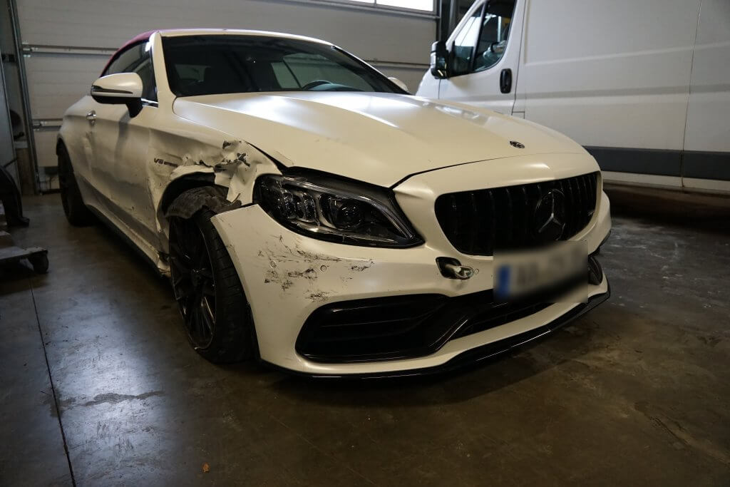 Ein weißer Mercedes mit starken Schäden an der linken Front steht in einer Halle neben einem weißen Transporter