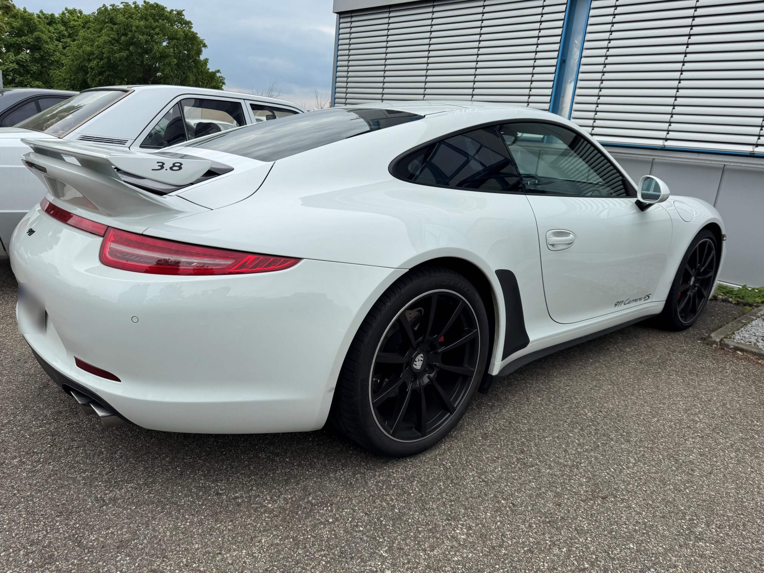 Ein weißer Porsche 911 Sportwagen mit Heckspoiler und schwarzen Felgen steht neben einem Oldtimer auf einer gepflasterten Fläche